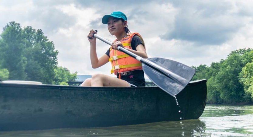 A young person wearing a life jacket paddles a canoe on calm water. There are trees in the background. 